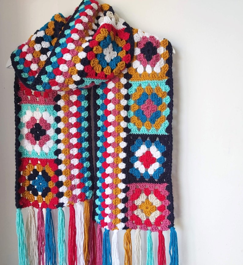 May include: A colorful crocheted scarf with a geometric pattern in shades of red, blue, green, yellow, and white. The scarf has a fringe on the bottom and is made of a soft, textured yarn.