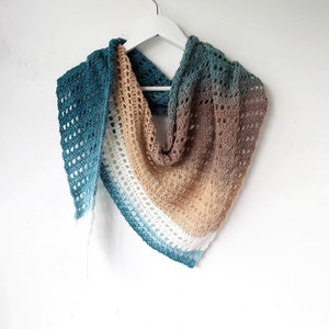 May include: A crocheted shawl with a gradient of teal, beige, white, and brown colors. The shawl is hanging on a white hanger against a white background.