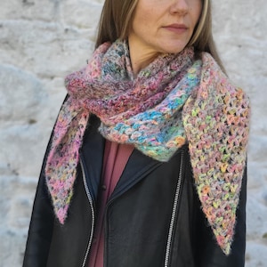 May include: A colorful, hand-crocheted shawl in a triangular shape. The shawl features a mix of pastel colors, including pink, blue, yellow, and purple. The open weave design adds texture and visual interest to the accessory.