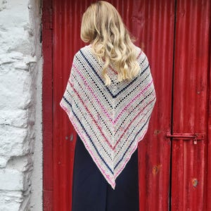 May include: A white crocheted shawl with colorful stripes and a geometric pattern. The shawl is worn by a person with long blonde hair.