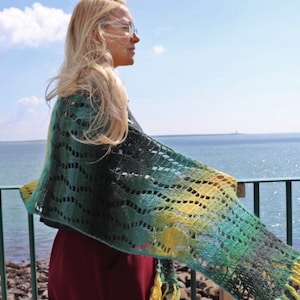 May include: A woman wearing a green, black, and yellow crocheted shawl with a fringe. She is standing on a deck overlooking the ocean.