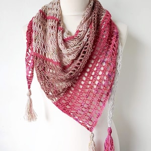 以下が含まれることがあります： マネキンに展示された、ピンク、ベージュ、グレーの色合いの手作りのオープンウィーブショール。 三角形のショールは、タッセル付きのエッジが特徴です。 色のグラデーションは、明るいベージュから濃いピンクへと変化し、視覚的に魅力的なアクセサリーを作り出しています。