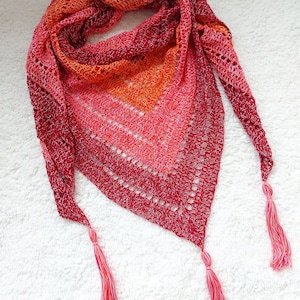 Puede incluir: Un chal de ganchillo con un degradado de colores rojo, naranja y amarillo. El chal tiene una forma triangular y presenta dos largas borlas.