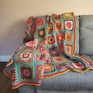 May include: A vibrant, hand-crocheted blanket in a patchwork design, draped over a grey sofa. The blanket features square motifs in teal, pink, yellow, orange, and brown. It has a bright yellow border around the edges.