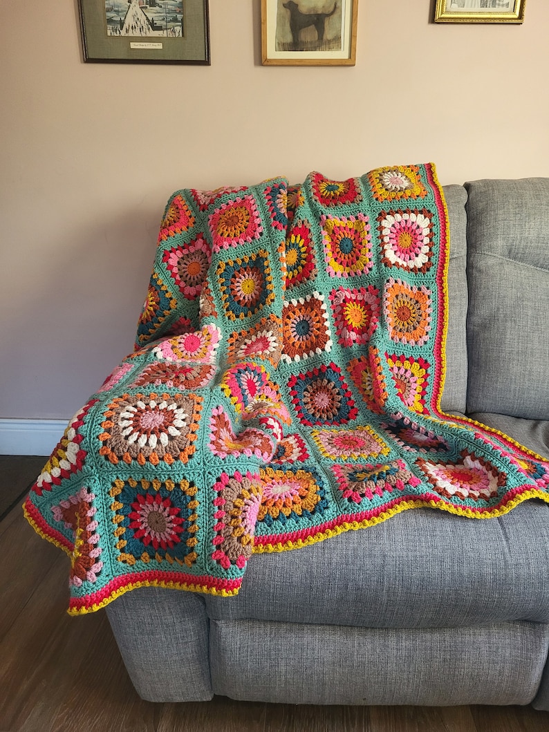 May include: A vibrant, hand-crocheted blanket in a patchwork design, draped over a grey sofa. The blanket features square motifs in teal, pink, yellow, orange, and brown. It has a bright yellow border around the edges.