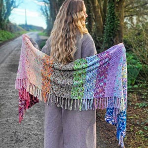 Pode incluir: Um xale colorido com franjas e um gradiente de cores, incluindo rosa, laranja, verde e roxo. O xale é segurado aberto, mostrando os tons vibrantes e o tecido texturizado. O fundo apresenta uma estrada ladeada por árvores.