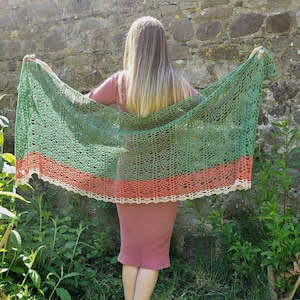 Könnte beinhalten: Ein gehäkelter Schal in Grün, Orange und Creme wird vor einer Steinmauer offen gehalten. Der Schal hat einen gewellten Rand und ein Blumenmuster. Die Person, die den Schal hält, trägt ein rosa Kleid.