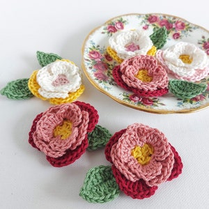 May include: Five crocheted flowers with green leaves. The flowers are in shades of pink, red, white, and yellow. The flowers are arranged on a white surface and a vintage floral patterned plate.
