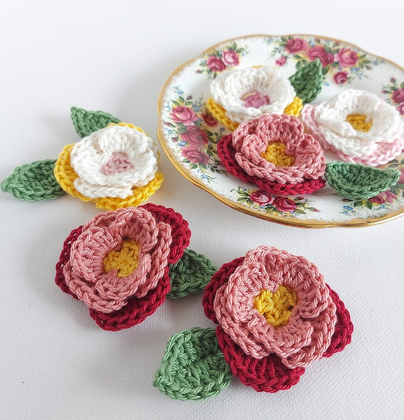 May include: Five crocheted flowers with green leaves. The flowers are in shades of pink, red, white, and yellow. The flowers are arranged on a white surface and a vintage floral patterned plate.