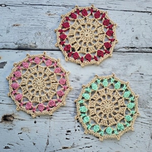 May include: Three circular crochet coasters featuring a beige base with decorative raised stitches in dark red, light pink, and mint green. Each coaster has a scalloped edge and a central star-like pattern created with intricate yarn loops.