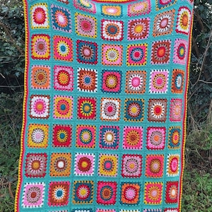 May include: A vibrant, hand-crocheted blanket. It has a turquoise base with many squares in shades of pink, yellow, orange, and brown. The edges are finished with a yellow and pink border. The blanket is shown outdoors, against a backdrop of greenery.