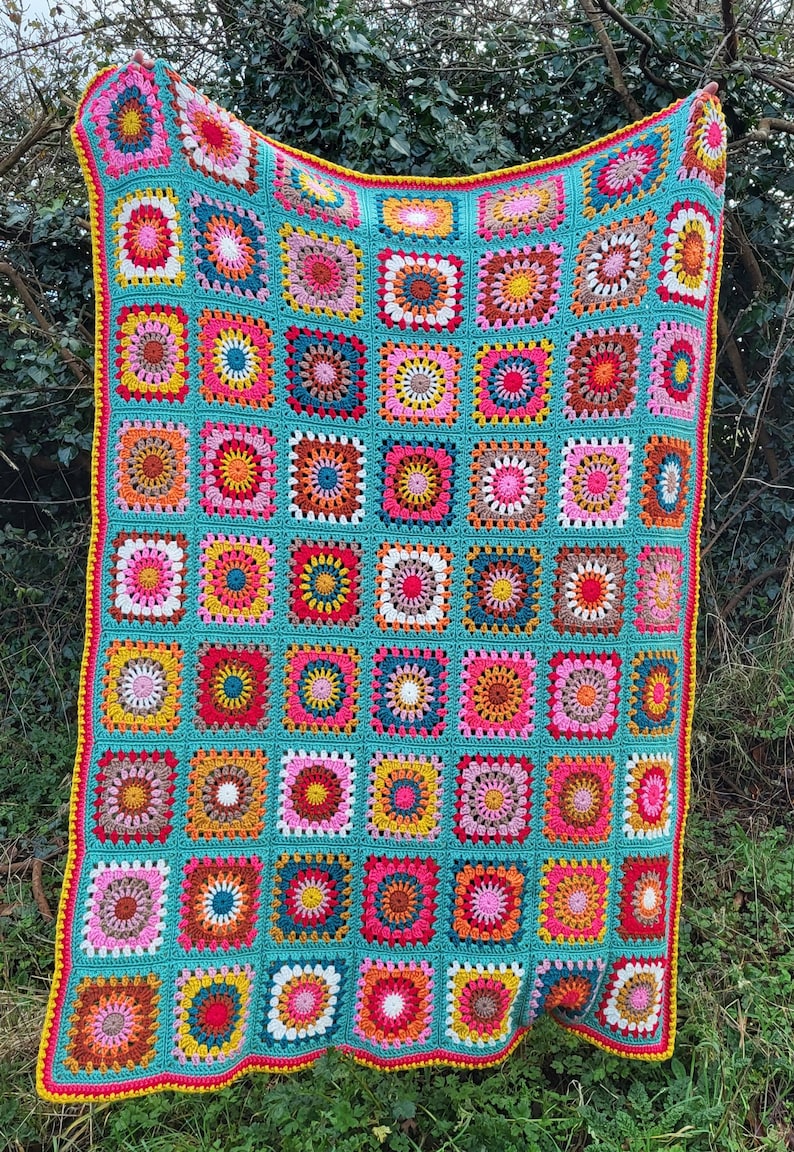 May include: A vibrant, hand-crocheted blanket. It has a turquoise base with many squares in shades of pink, yellow, orange, and brown. The edges are finished with a yellow and pink border. The blanket is shown outdoors, against a backdrop of greenery.