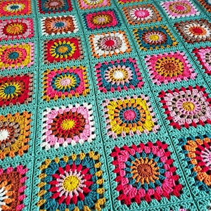 May include: A vibrant, hand-crocheted blanket with a patchwork design. The blanket is composed of square motifs in shades of pink, yellow, orange, and teal. Each square features a floral pattern, and the blanket is edged with teal yarn.