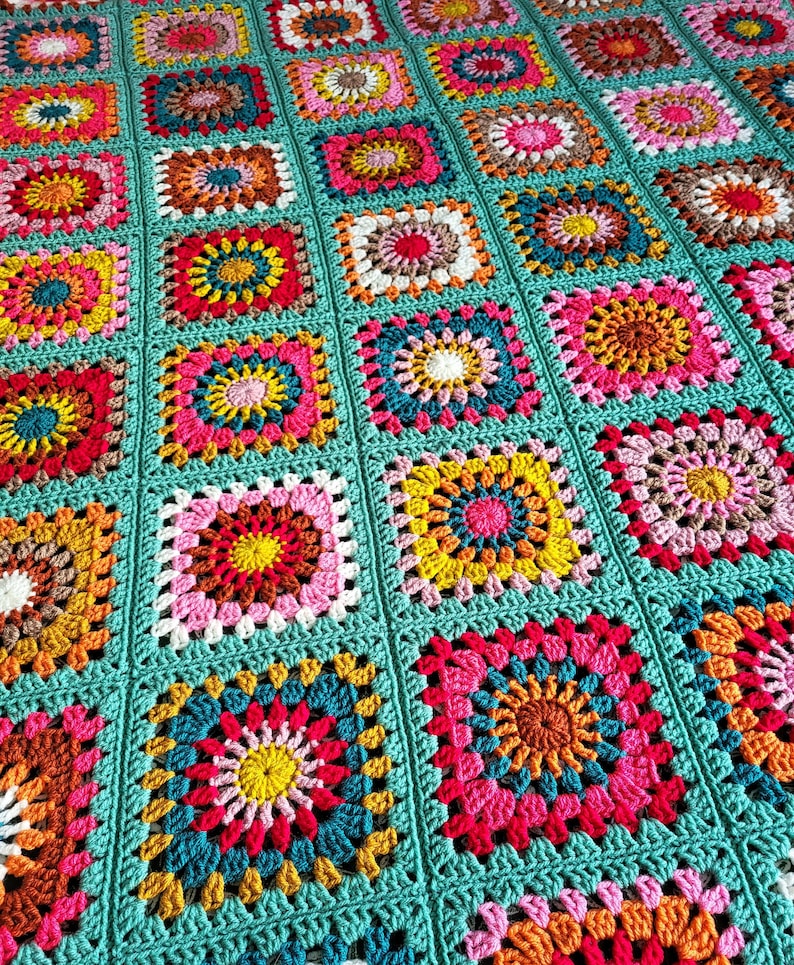 May include: A vibrant, hand-crocheted blanket with a patchwork design. The blanket is composed of square motifs in shades of pink, yellow, orange, and teal. Each square features a floral pattern, and the blanket is edged with teal yarn.
