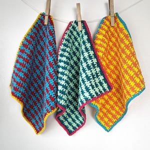 May include: Three crocheted dishcloths in different colors and patterns. The dishcloths are hanging on a clothesline. The dishcloths are made with a striped pattern in blue, red, and yellow.