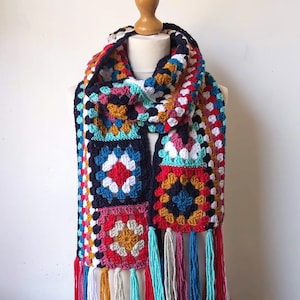 May include: A colorful crocheted scarf with a geometric pattern and fringe. The scarf is made with a variety of colors, including blue, red, white, yellow, and green.