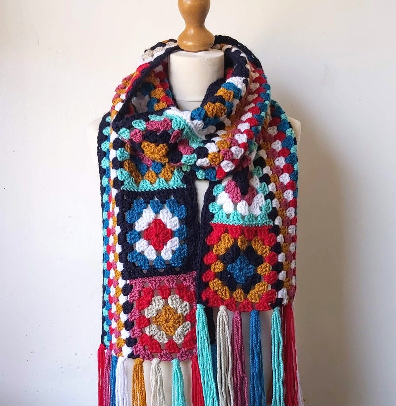 May include: A colorful crocheted scarf with a geometric pattern and fringe. The scarf is made with a variety of colors, including blue, red, white, yellow, and green.