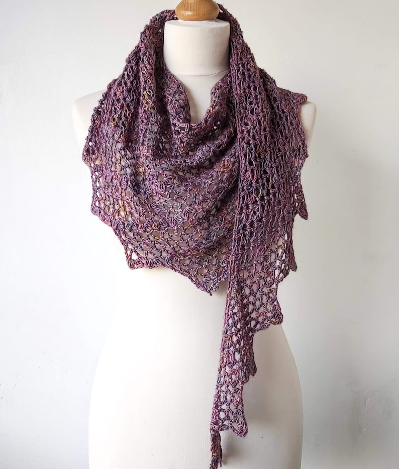 Puede incluir: Un chal de ganchillo morado y marr&oacute;n con un dise&ntilde;o de encaje. El chal est&aacute; drapeado sobre un maniqu&iacute; blanco.