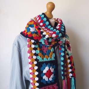 May include: A colorful crocheted scarf with a granny square pattern in shades of red, white, blue, green, and yellow. The scarf has long tassels on the ends.