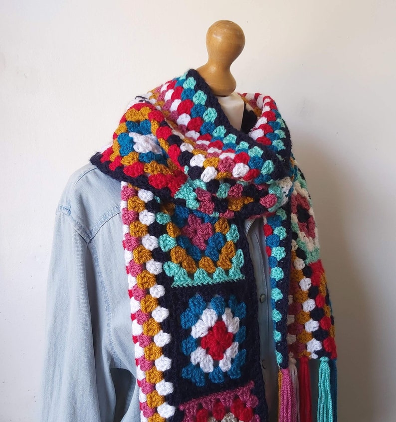 May include: A colorful crocheted scarf with a granny square pattern in shades of red, white, blue, green, and yellow. The scarf has long tassels on the ends.