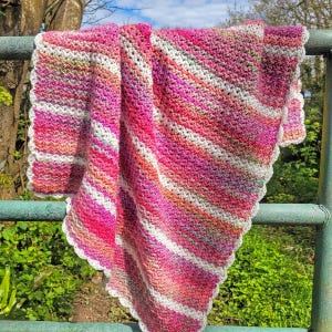 May include: A handmade crochet blanket in shades of pink, white, and orange, with a scalloped edge. The blanket is draped over a green metal railing, showcasing its diagonal striped pattern. The yarn appears soft and textured, perfect for a cozy home decor item.