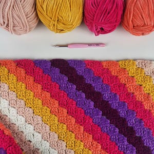 May include: A colorful crocheted blanket with a diagonal pattern in shades of pink, orange, yellow, red, purple, and white.