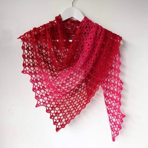 May include: A red and pink crocheted shawl with a lacy pattern. The shawl is draped over a white hanger.