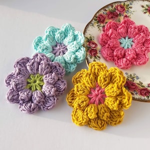 May include: Four crocheted flower embellishments in different colors: purple, teal, yellow, and pink. The flowers are made with a textured stitch and have a center circle.