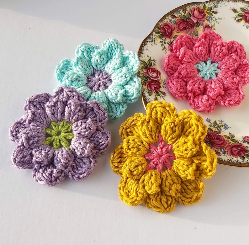 May include: Four crocheted flower embellishments in different colors: purple, teal, yellow, and pink. The flowers are made with a textured stitch and have a center circle.