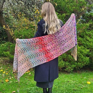 May include: A colorful crocheted shawl with a scalloped edge. The shawl is made with a variety of colors, including pink, purple, blue, and red. The shawl is draped over the shoulders of a person standing in a garden.