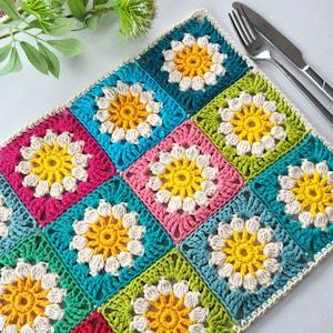 May include: A colourful crocheted placemat with a granny square pattern in shades of pink, green, blue, and yellow. The placemat is made with white yarn and has a scalloped edge.