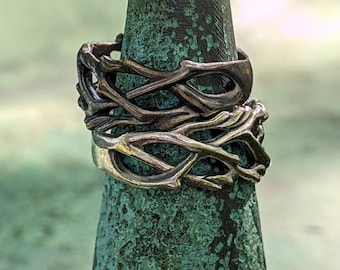Könnte beinhalten: Zwei silberne Ringe mit einem zweigartigen Design, die übereinander gestapelt sind. Die Ringe werden auf einem dunkelgrünen, kegelförmigen Objekt präsentiert. Der Hintergrund ist ein verschwommenes Grün.