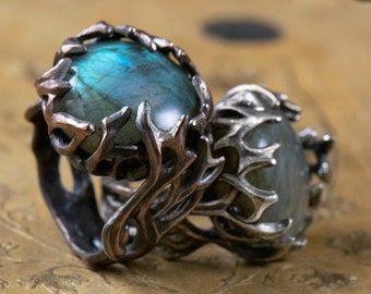 Puede incluir: Dos anillos de plata con grandes piedras preciosas ovaladas e iridiscentes. Los anillos tienen un diseño orgánico detallado, con el metal que parece envolver las piedras. Las piedras preciosas tienen una mezcla de tonos azules, verdes y grises. Los anillos están sobre una superficie dorada.