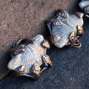 May include: Three silver and bronze metal charms shaped like seashells with intricate details. Each charm has a different design and is attached to a loop.