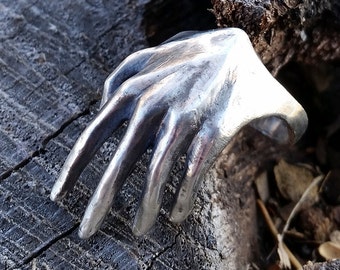 May include: A silver ring shaped like a hand reaching out, resting on a piece of weathered wood.