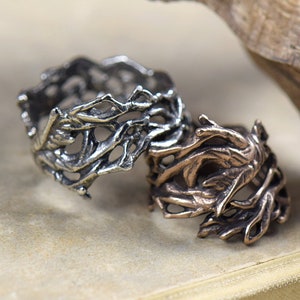 Könnte beinhalten: Zwei Ringe aus Metall, einer silber und einer bronzefarben, sind in Form von Ästen eines Baumes gestaltet. Die Ringe liegen auf einer hellbraunen Oberfläche.