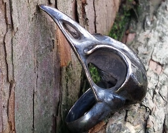 Puede incluir: Un anillo metálico oscuro con forma de cráneo de ave, con un pico y una cuenca ocular detallados. El anillo está colocado sobre un fondo de tronco de árbol marrón texturizado. La palabra "HIBERNACULA" es visible en la parte inferior.