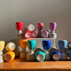 May include: A collection of 15 colourful rubber stamps with different designs. The stamps are arranged in three rows on a wooden surface. The stamps are all different colours, including red, blue, green, yellow, and purple. Some of the stamps have a floral design, while others have a geometric design.