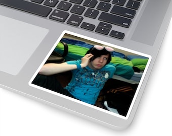May include: A young person with black hair and pink glasses is on a phone call. They are wearing a blue shirt with a graphic design and are sitting on a bed with green and blue bedding.