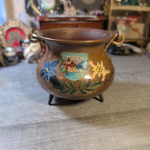 May include: A small, copper-colored pot with three legs and two handles. The pot is decorated with hand-painted floral and landscape designs, including a blue flower, a shield with a castle scene, and a yellow flower. The pot has a rustic, vintage aesthetic.
