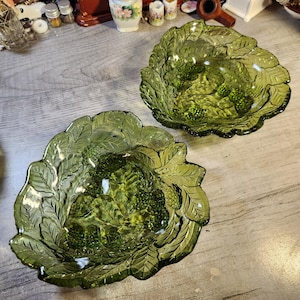 May include: Two green glass bowls with a leaf and berry design. The bowls are made of clear glass with a green tint and have a raised design of leaves and berries.