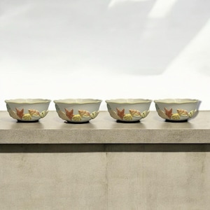 May include: Four small, off-white ceramic bowls with a scalloped rim. Each bowl is decorated with a repeating pattern of orange, yellow, and green leaf designs. The bowls are arranged in a row on a light-colored surface.