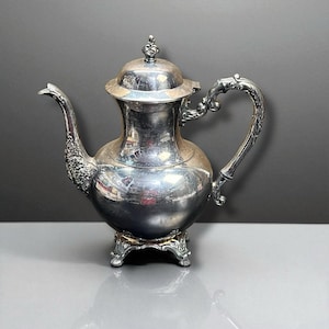 May include: A silver-colored antique teapot with ornate detailing. The teapot features a curved spout, a decorative handle, and a lid with a small finial. It sits on a decorative base, reflecting light and adding to its vintage charm.