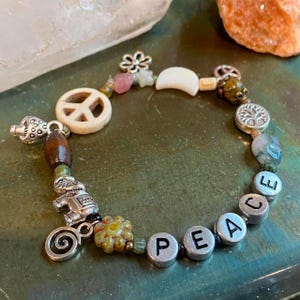 Puede incluir: Una pulsera de cuentas con un dije de símbolo de la paz, cuentas de madera y dijes plateados. La pulsera deletrea "PEACE" con cuentas de letras plateadas. Otras cuentas incluyen acentos rosas, verdes y blancos. La pulsera está sobre una superficie verde.