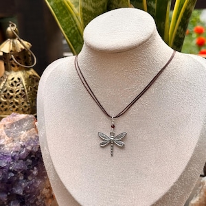 May include: A silver dragonfly pendant necklace with a brown cord. The dragonfly has detailed wings and a small red bead above it. The necklace is displayed on a mannequin.