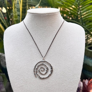 May include: A silver pendant necklace with a spiral design. The pendant is suspended from a dark brown cord. The necklace is shown on a white mannequin bust, set against a background of green leaves.