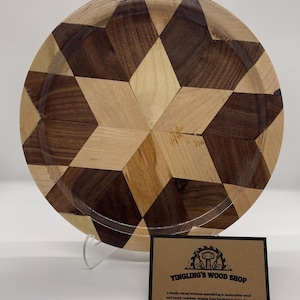 May include: A handcrafted wooden plate with a geometric starburst design. The plate features alternating dark and light wood pieces, creating a striking visual pattern. The plate is displayed on a clear stand, with a business card from Yingling's Wood Shop.