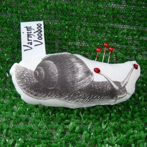 May include: A white fabric snail pincushion with a black and white illustration of a snail. The pincushion has red plastic push pins sticking out of it. The pincushion has a white tag attached to it that says "Varmint Voodoo".