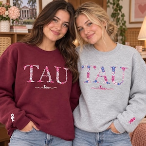 May include: A maroon sweatshirt and a grey sweatshirt, each featuring the word TAU in a floral pattern on the chest, the word tau in cursive below, and a small ribbon symbol embroidered on the sleeve cuff, made of a soft cotton blend fabric.