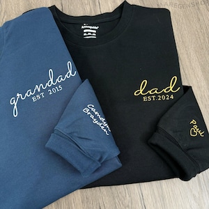Peut inclure: Deux t-shirts à manches longues, un bleu marine et un noir. Le t-shirt bleu marine porte l'inscription "grandad EST 2015" en écriture blanche. Le t-shirt noir porte l'inscription "dad EST.2024" en écriture dorée. La manche du t-shirt bleu marine porte l'inscription "Camdyn Brayden" et la manche du t-shirt noir porte l'inscription "Paul" avec un cœur.